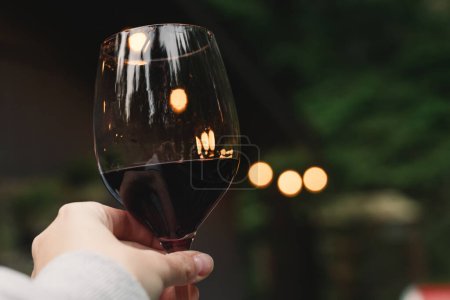 Photo for Woman holding a glass with red with blurred lights on the background, relax on the patio, cabin life - Royalty Free Image