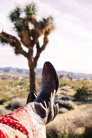 Nahaufnahme einer Frau in Cowboystiefeln mit Joshuabaum im Hintergrund, Boho-Lifestyle