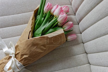 Photo for Bouquet of fresh pastel pink tulips wrapped in the craft paper decorated with satin ribbon lying on the beige chair, - Royalty Free Image