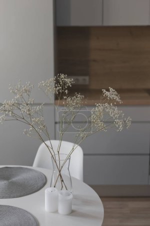 Téléchargez les photos : Table ronde blanche dans la salle à manger en face de la cuisine scandinave en bois et gris, design nordique élégant, fleurs dans un vase en verre et décoration intérieure. - en image libre de droit