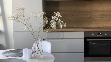 Mesa redonda blanca en comedor frente a cocina escandinava de madera y gris, elegante diseño nórdico, flores en jarrón de vidrio y decoración interior.