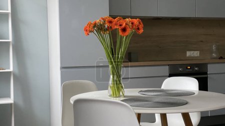 Téléchargez les photos : Table ronde blanche dans la salle à manger en face de la cuisine scandinave en bois et gris, design nordique élégant, fleurs dans un vase en verre et décoration intérieure. - en image libre de droit