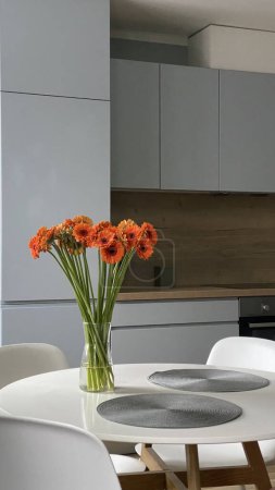 Mesa redonda blanca en comedor frente a cocina escandinava de madera y gris, elegante diseño nórdico, flores en jarrón de vidrio y decoración interior.