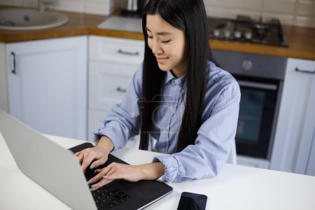 Photo for Freelance BIPOC programmer coding on notebook computer. Cheerful Vietnamese female person typing text on laptop keyboard in home kitchen - Royalty Free Image