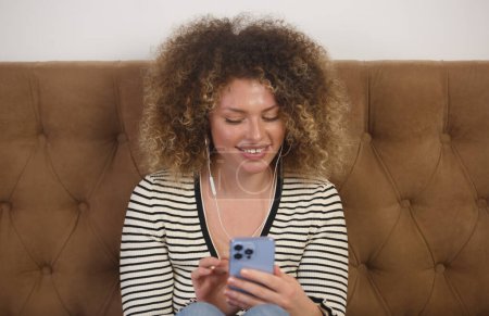 Téléchargez les photos : Happy young woman enjoying music in headphones. Cheerful curly female person sitting in bedroom and listening to favorite musical tracks on the phone - en image libre de droit