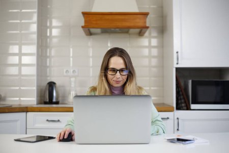 Photo for Adult freelancer woman works on computer in home kitchen. Pretty lady in 40s doing distant work online with modern laptop - Royalty Free Image