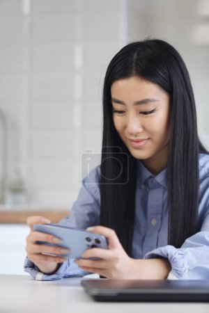 Téléchargez les photos : Happy asian girl plays online game on smart phone. Cheerful young Vietnamese female person playing games on modern gadget at home - en image libre de droit