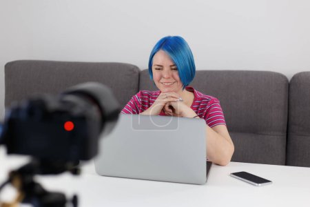 Foto de Cheerful video blogger woman with dyed blue hair talks on professional camera on tripod. Online coach films blog video at home - Imagen libre de derechos