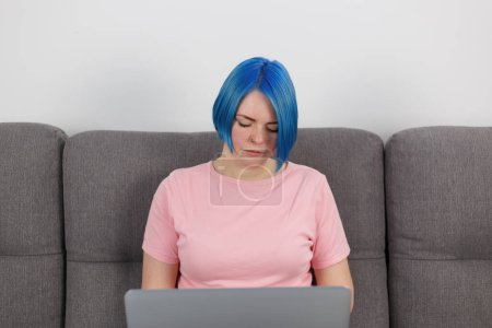 Téléchargez les photos : Millennial woman with dyed blue hair works on notebook computer on couch at home. Portrait of focused freelancer person working on laptop - en image libre de droit