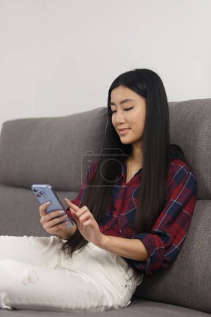 Téléchargez les photos : Beautiful asian girl browsing mobile app on smart phone while sitting on sofa at home. Young Vietnames BIPOC female using mobile phone - en image libre de droit