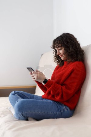 Foto de Young girl sitting on couch and reading ebook. Curly white female person in early 20s browsing internet on tablet computer device - Imagen libre de derechos