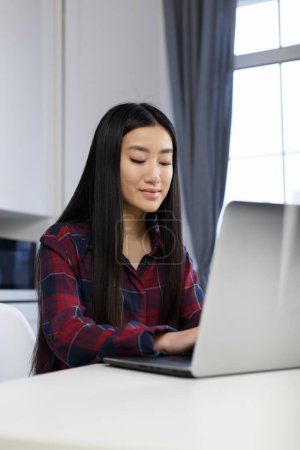 Téléchargez les photos : Belle fille vietnamienne utilisant un ordinateur portable pour étudier en ligne. Jeune femme asiatique travaille sur ordinateur portable moderne à la maison avec un sourire joyeux - en image libre de droit
