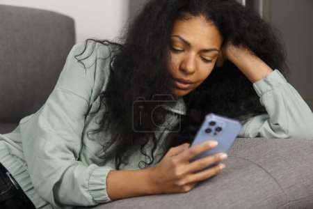 Photo for Young adult black woman lying on couch and using mobile phone app. Portrait of BIPOC female browsing internet on smart phone - Royalty Free Image