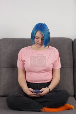 Téléchargez les photos : Jeune femme aux cheveux bleus teints assise sur le canapé à la maison et tapant un message dans un téléphone intelligent - en image libre de droit
