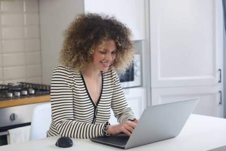 Photo for Happy white woman with curly hair working on silver laptop computer at home. Cheerful young female using modern notebook pc with a smile - Royalty Free Image