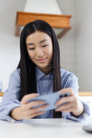 Téléchargez les photos : Belle fille asiatique joue jeu en ligne avec un téléphone intelligent. Gai Vietnamien POC femelle en utilisant un téléphone bleu moderne pour jouer à des jeux vidéo - en image libre de droit