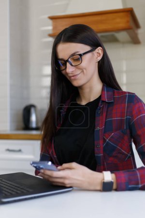 Photo for Cheerful brunette female person using modern smart phone at home. Young adult woman in glasses browsing mobile app on cellphone - Royalty Free Image
