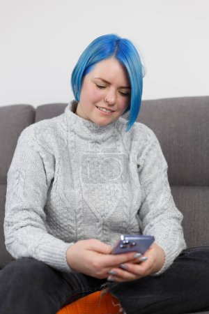 Téléchargez les photos : Jeune femme adulte aux cheveux bleu vif assise sur le canapé et tapant un message sur un téléphone intelligent. Femme blanche gaie utilisant un téléphone portable bleu moderne pour la communication en ligne - en image libre de droit