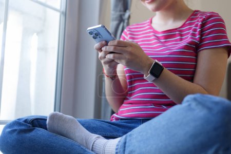 Téléchargez les photos : Jeune fille assise à la maison et tapant un message sur un téléphone intelligent. Femme blanche méconnaissable utilisant un téléphone portable moderne pour la communication - en image libre de droit