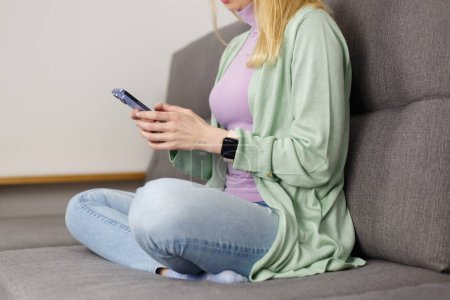 Photo for Adult woman sitting on couch and typing a message on smart phone. Unrecognizable white female person using modern mobile phone app at home - Royalty Free Image