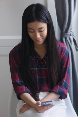 Téléchargez les photos : Belle fille asiatique naviguant téléphone mobile à la maison. Portrait de jolies jeunes adultes POC de Xoname en utilisant un téléphone intelligent moderne - en image libre de droit
