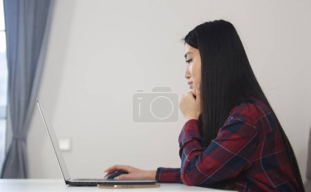 Photo for Young adult Asian female studying online. Beautiful Vietnamese girl working on laptop computer at home, looking at screen in thought - Royalty Free Image