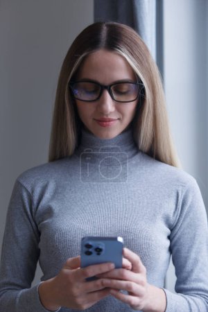 Foto de Beautiful blonde woman in glasses typing message on mobile phone. Portrait of attractive white female standing by the window at home and texting online with a smart phone - Imagen libre de derechos