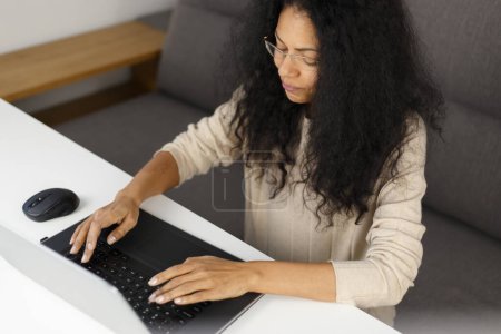 Photo for Young adult black woman working on notebook computer at home. Overhead photo of natural looking POC female typing text on laptop keyboard - Royalty Free Image