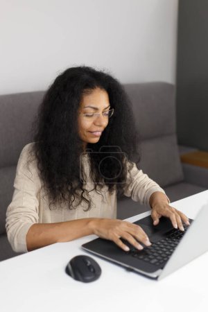 Téléchargez les photos : Femme noire joyeuse dans des lunettes assis derrière le bureau et tapant du texte sur l'ordinateur. Jeune adulte POC femelle travaillant sur ordinateur portable moderne à la maison - en image libre de droit