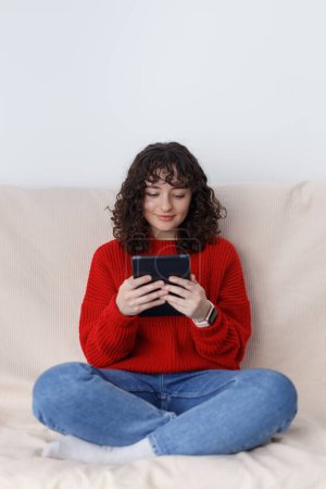 Foto de Niña sentada en el sofá y leyendo las noticias de la tableta. Alegre hembra blanca usando gadget portátil moderno en casa - Imagen libre de derechos
