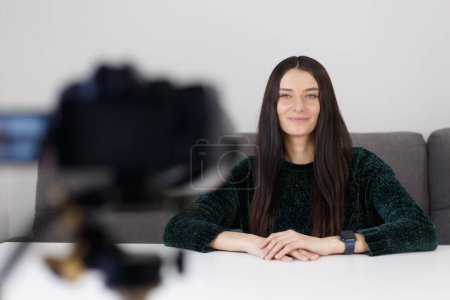 Foto de Beautiful influencer woman talking on videocamera at home. Cheerful young adult female person sitting at home and filming video blog with a camcorder on a tripod - Imagen libre de derechos