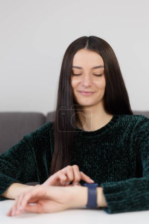 Foto de Alegre joven mujer que navega notificaciones en el reloj inteligente. Retrato de una hermosa mujer blanca sentada detrás de un escritorio en casa y utilizando relojes de pulsera inteligentes modernos - Imagen libre de derechos