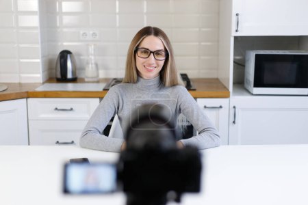 Foto de Cheerful blonde woman in glasses filming a video blog in home kitchen. Friendly white female person in 30s speaking on camcorder camera - Imagen libre de derechos