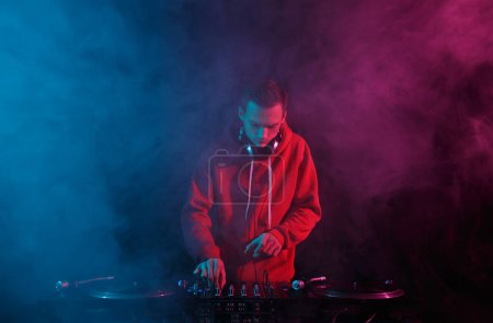 Téléchargez les photos : Disc jockey mixing hip hop music with vinyl records and sound mixer in smoke on stage. Night club DJ plays set with turntables - en image libre de droit
