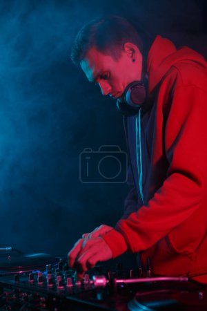 Téléchargez les photos : Cool jeune DJ en sweat à capuche rouge joue de la musique sur la fête en fumée. Homme blanc jouant ensemble dans la boîte de nuit - en image libre de droit