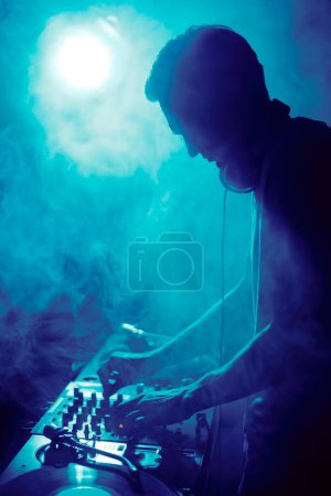 Téléchargez les photos : Silhouette de DJ de club jouant de la musique sur techno party en boîte de nuit. Disc jockey mixant des morceaux musicaux sur scène en fumée - en image libre de droit