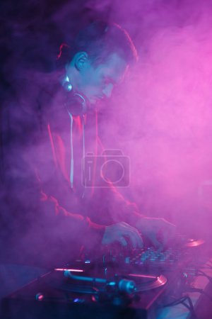 Foto de El DJ del club nocturno toca en la fiesta de hip hop. Disco jockey mezclando pistas musicales con dispositivo mezclador de sonido en humo - Imagen libre de derechos