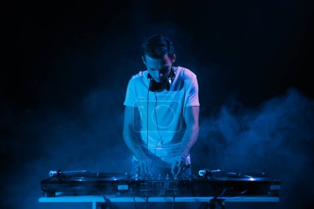 Foto de Club DJ toca música con humo y luces azules. Disco jockey mezcla de pistas musicales en el escenario del concierto en discoteca - Imagen libre de derechos