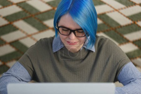 Téléchargez les photos : Portrait de femme blanche aux cheveux bleus teints travaillant sur ordinateur à la maison. Photo rapprochée de la femme dans des lunettes étudiant en ligne avec un ordinateur portable - en image libre de droit