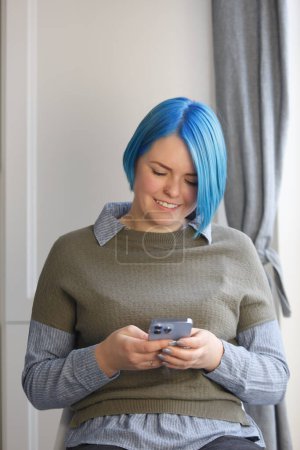 Téléchargez les photos : Joyeux millenial femelle avec les cheveux bleus teints assis sur une chaise par la fenêtre et dactylographier sms message sur téléphone intelligent - en image libre de droit