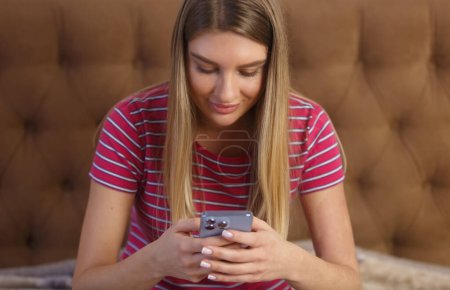 Téléchargez les photos : Cheerful white female sitting on bed and typing a message on a smart phone. Portrait of beautiful young woman using modern mobile phone in bedroom - en image libre de droit
