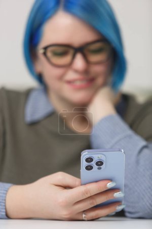 Téléchargez les photos : Young woman holding modern smart phone in hand. Cheerful white female with blue hair browing mobile app on a blue phone. Focus on hand with gadget - en image libre de droit