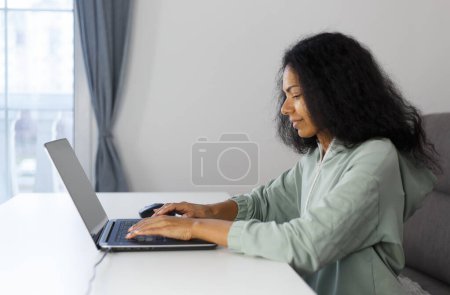 Photo for Freelance programmer coding on a laptop at home. Focused black woman typing text on modern computer with a smile - Royalty Free Image