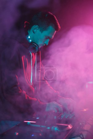 Téléchargez les photos : Club DJ mélangeant musique en fumée et néons. Hip hop disc jockey jouant des morceaux musicaux sur la fête dans une boîte de nuit - en image libre de droit