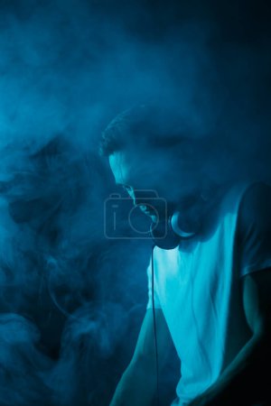 Téléchargez les photos : DJ playing music on techno party in smoke and blue neon lights. Young disc jockey plays set in nightclub - en image libre de droit
