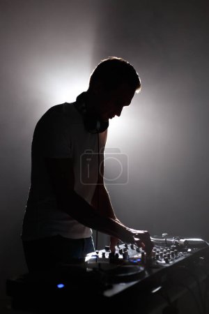 Téléchargez les photos : Silhouette of club dj mixing techno music. Young white man perfroming as a disc jockey on party in dark night club - en image libre de droit