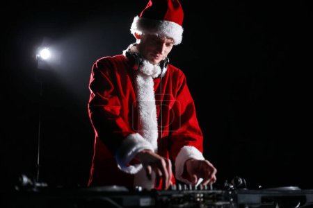 Téléchargez les photos : Santa DJ jouant de la musique à la fête de Noël dans une boîte de nuit. Disc jockey portant traditionnelle tenue de Noël rouge joue mis sur scène - en image libre de droit