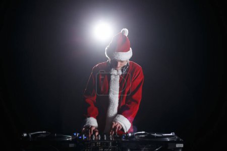 Photo for Christmas party DJ in Santa costume plays music on stage. Disc jockey wearing traditional red Xmas outfit playing set on party in night club - Royalty Free Image