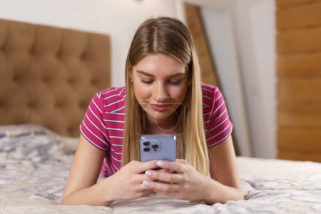 Téléchargez les photos : Beautiful blonde girl lying on bed in bedroom and typing a message on smart phone. Portrait of pretty white woman in 20s using modern mobile phone for communication - en image libre de droit