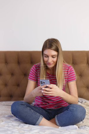 Téléchargez les photos : Beautiful young girl with blonde hair sitting on bed in bedroom and typing sms on mobile phone. Pretty white female texting online with modern smart phone - en image libre de droit